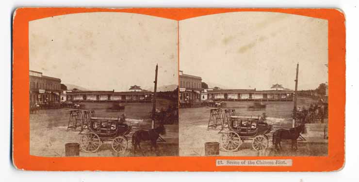 This stereograph shows a stagecoach in front of a square lined with adobe buildings.